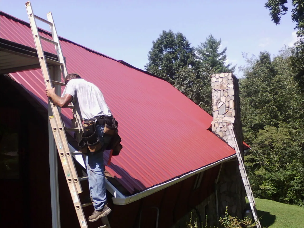 Roofer on a ladder beside a red metal roof and stone chimney — Soffit & Fascia Repair by Roofing Brothers in Joseph City